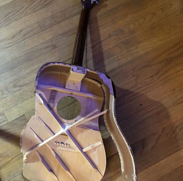 Broken acoustic guitar with torn top and exposed bracing on hardwood floor, next-level jerks