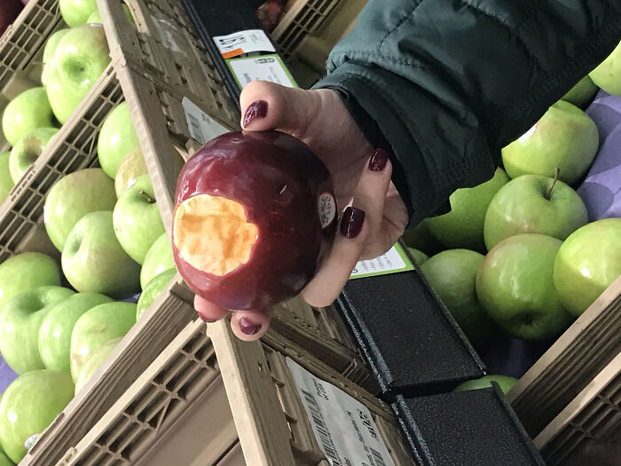 Hand holding a red apple with a large bite in supermarket produce section, next-level jerks