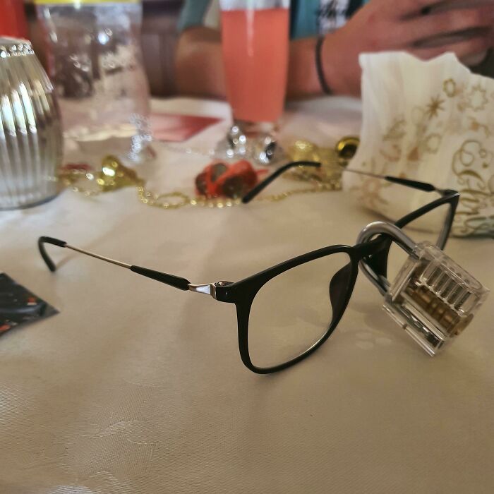 Black-framed eyeglasses on a table with a transparent padlock attached, with drinks and decorations blurred in the background.