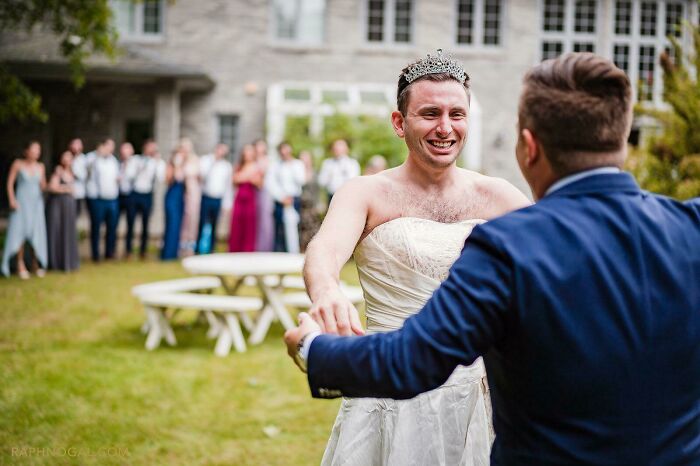 Man wearing a tiara and wedding dress, smiling and holding hands with a sibling in a joyful outdoor moment.
