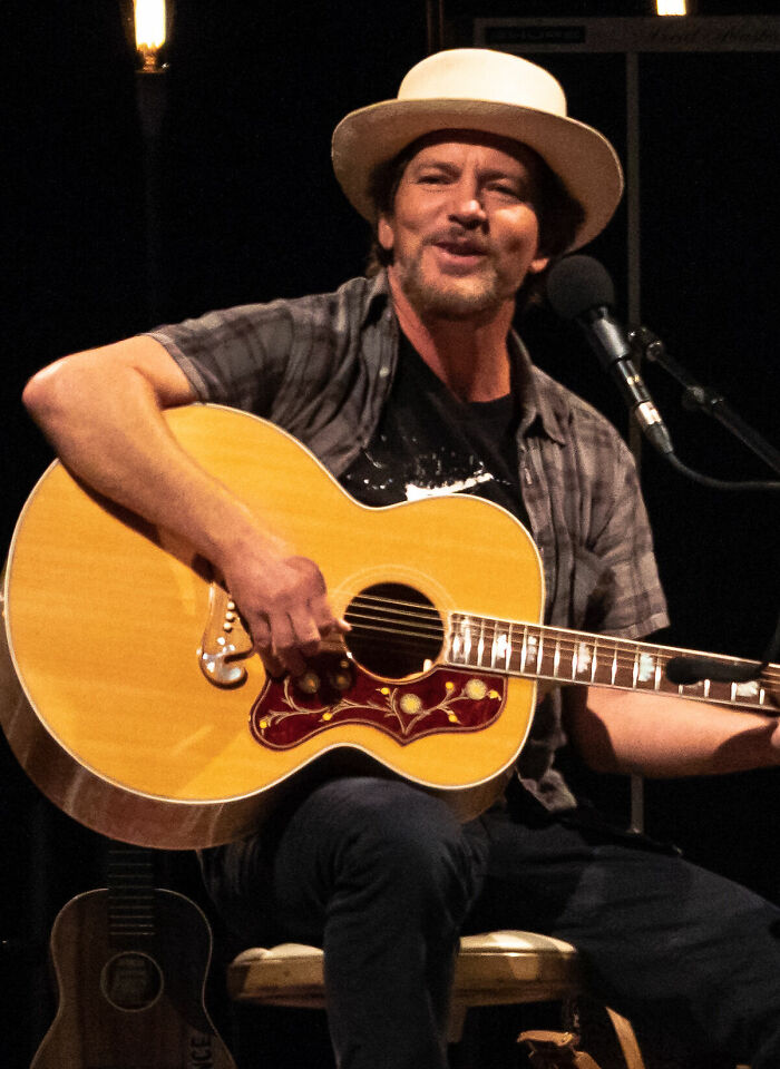 Eddie Vedder playing acoustic guitar, wearing a hat and plaid shirt during a live musical performance.