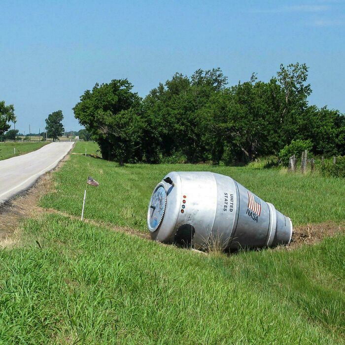 NASA space capsule replica resting in a grassy roadside ditch beside a small American flag and trees in daylight