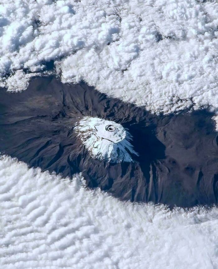 A volcanic mountain peak covered in snow, seen from above through a break in the surrounding clouds.