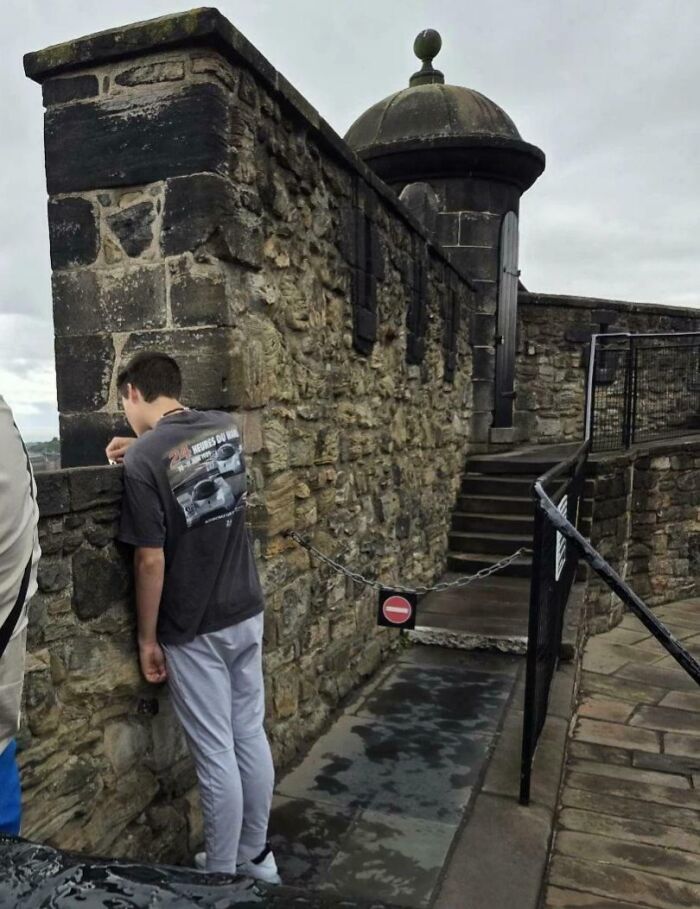 Young man leaning over stone castle wall by no-entry chain, next-level jerks behavior