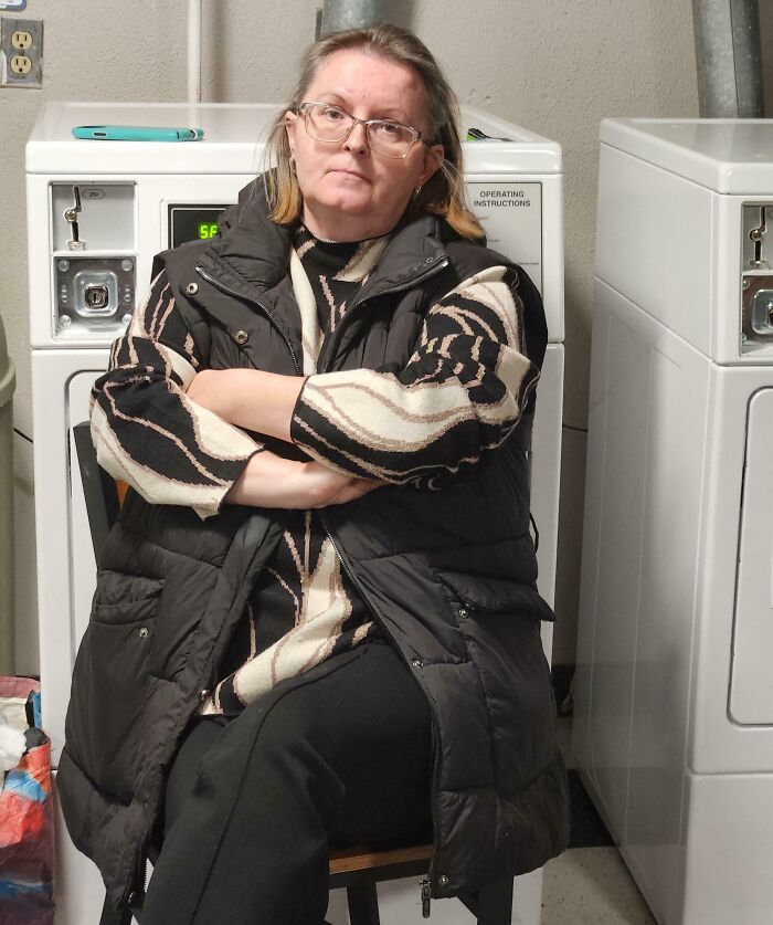 Middle-aged woman sitting with arms crossed by laundry machines, annoyed expression, an example of next-level jerks