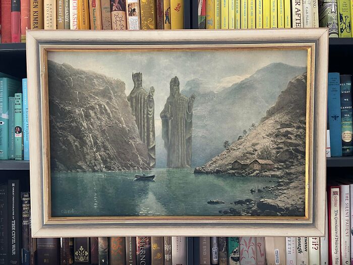 Framed thrifted painting showing a lake with large humorous statues edited into a mountainous landscape.