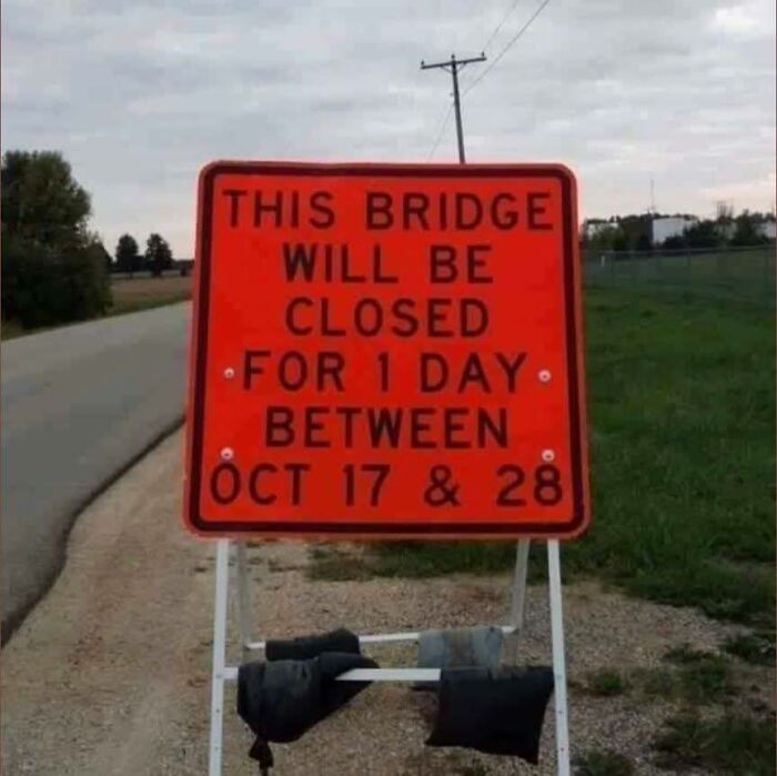 Orange funny stupid sign showing confusing road closure dates on a rural roadside under a cloudy sky.