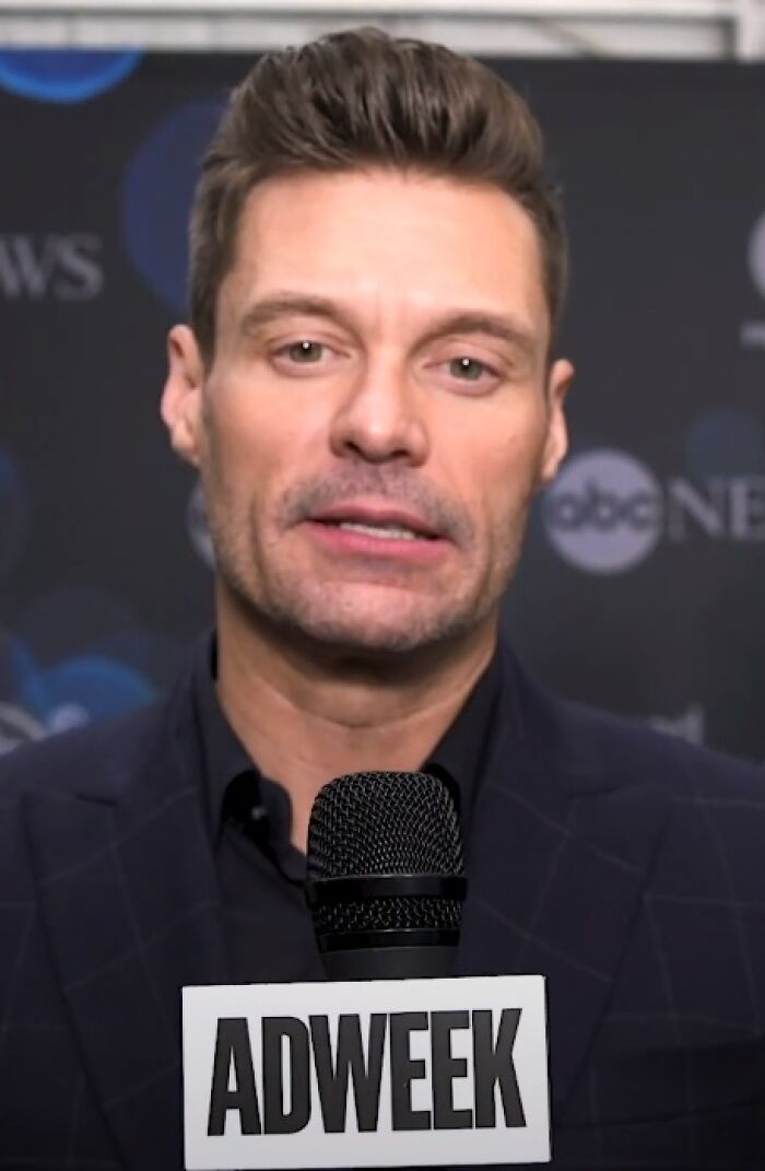Ryan Seacrest speaking at an event holding a microphone with Adweek logo, wearing a dark blazer and shirt.