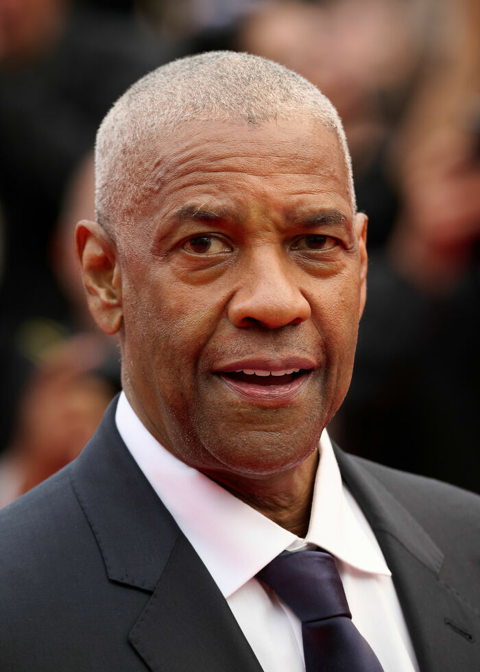Denzel Washington in a black suit and tie at a public event, highlighting his bio and career achievements.