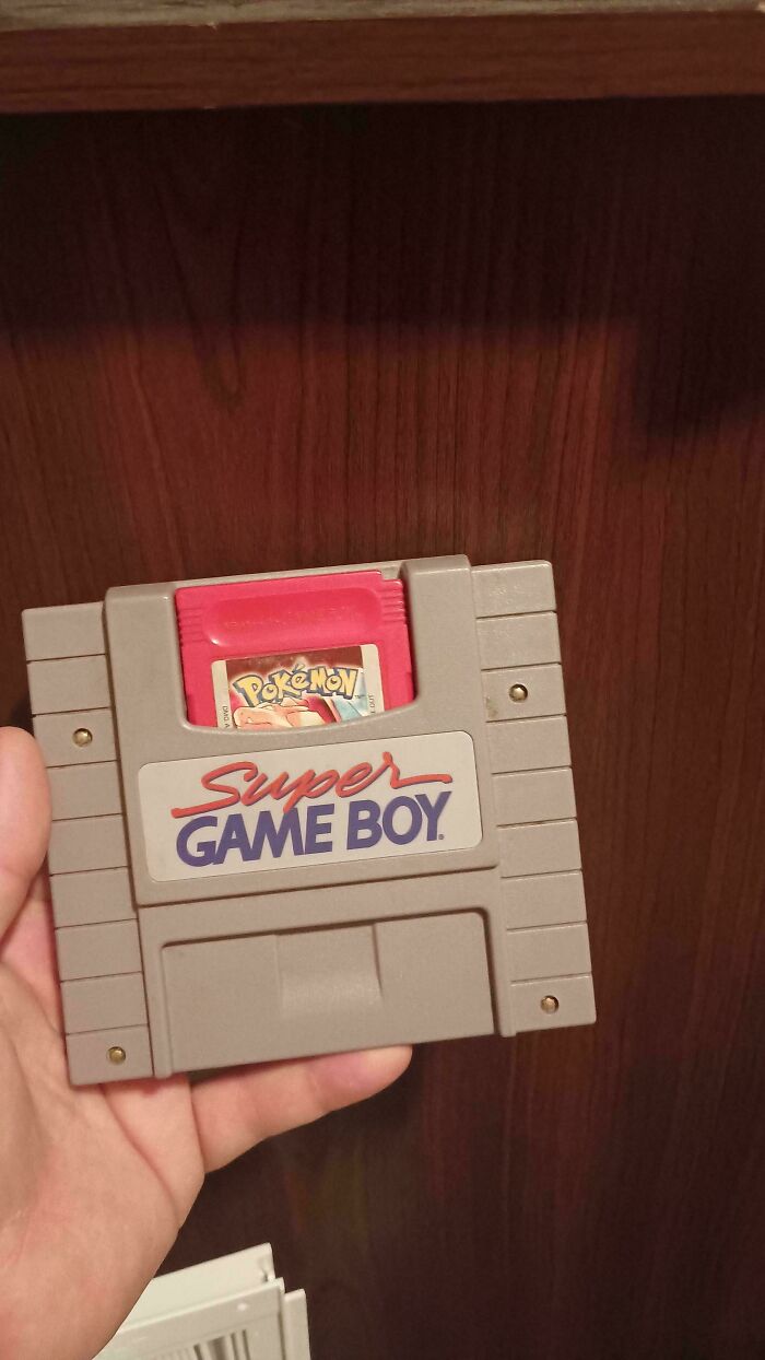 Hand holding a Super Game Boy cartridge with a red Pokémon game inserted, evoking 90s and 2000s nostalgia.