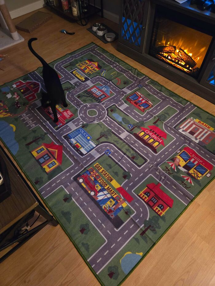 Black cat walking on a colorful play rug featuring roads and buildings, evoking 90s and 2000s nostalgia in a cozy room.