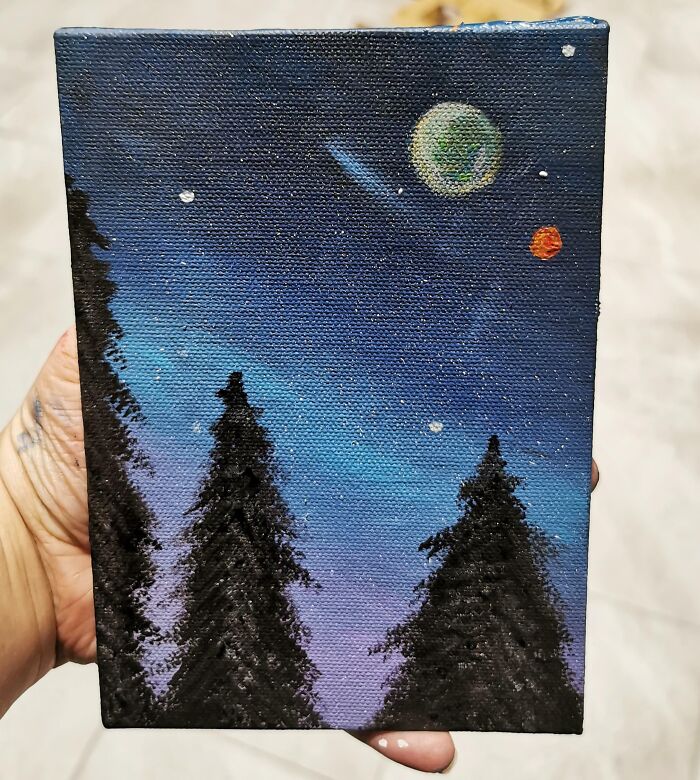 Hand holding a painting of silhouetted pine trees under a starry night sky with planets, capturing holiday moments so sweet.