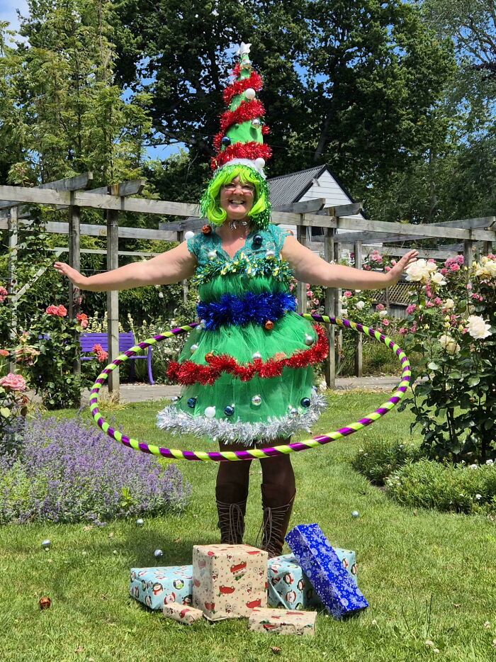 Woman dressed as a festive Christmas tree hula hooping outdoors with wrapped holiday presents around her feet.