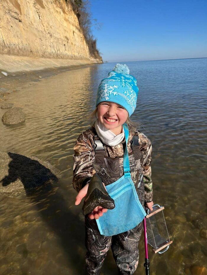 Smiling child in winter hat holding a large fossil by the water, capturing a sweet holiday moment outdoors.