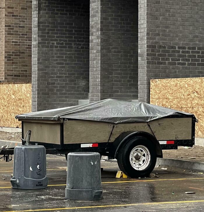 Remolque cubierto con lona en estacionamiento junto a dos botes de basura grises en imagen divertida de 2025.