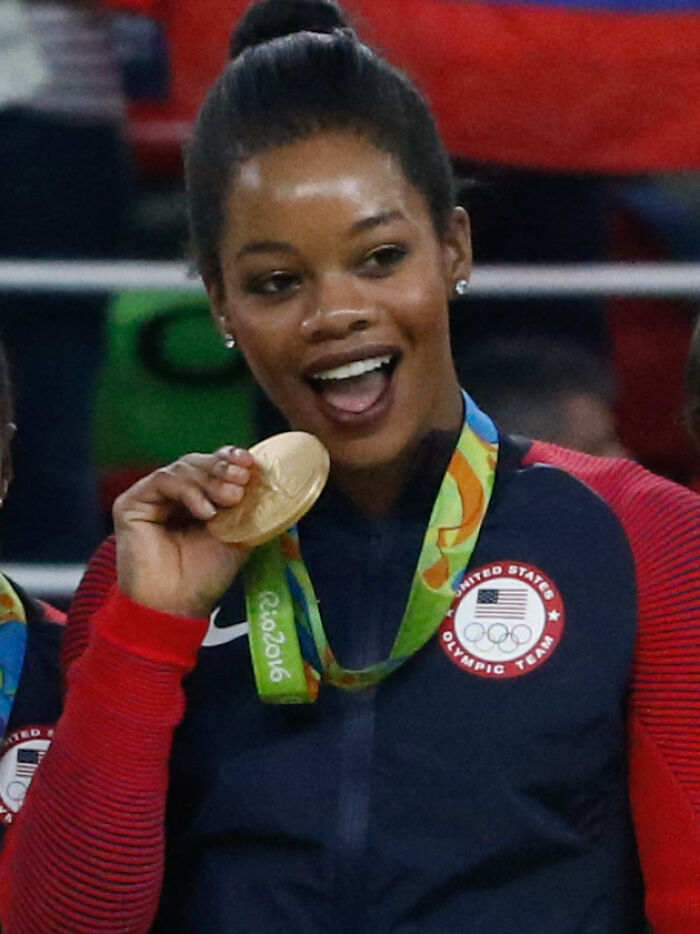 Gabby Douglas smiling and holding a gold medal wearing a United States Olympic Team jacket at Rio 2016 Olympics.