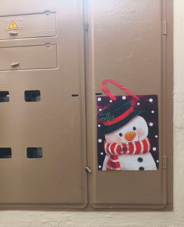 Snowman decoration with a red scarf and black hat attached to a plain brown electrical panel in a Lithuanian apartment building.