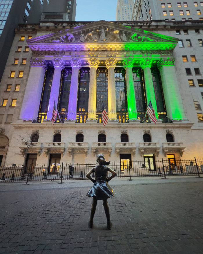 Fearless Girl statue facing the New York Stock Exchange building lit up in colorful lights in NYC.