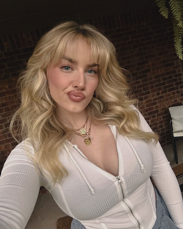 Young woman with blonde hair in a white zip-up top posing indoors, related to internet divided over seat switch on flight.