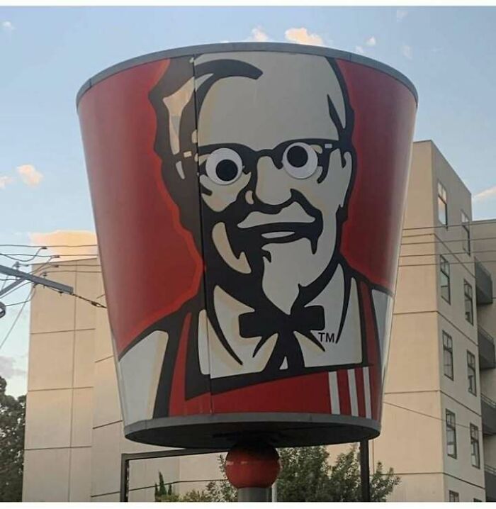 Large KFC bucket sign with cartoon Colonel Sanders wearing funny googly eyes in an urban setting, funny pics concept.