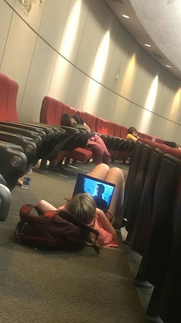 Person lying on floor between theater seats watching laptop, surrounded by others napping in a random funny moment.