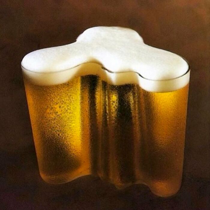 Trippy picture of a uniquely shaped glass filled with beer and topped with frothy foam on a dark background.