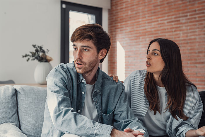 Man and woman in tense conversation on couch, man looking away worried about relationship and career pressure.