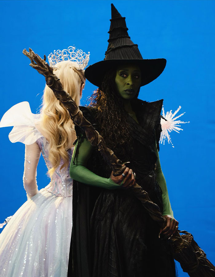 Cynthia Erivo as a green-skinned witch holding a staff, with a fairy-tale princess in a white dress behind her. Cynthia Erivo as a green-skinned witch holding a staff, with a fairy-tale princess in a white dress behind her.