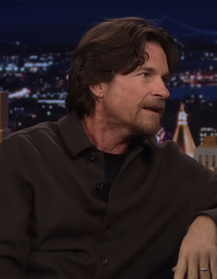 Jason Bateman in a dark shirt sharing a story during a nighttime talk show with city lights blurred in the background