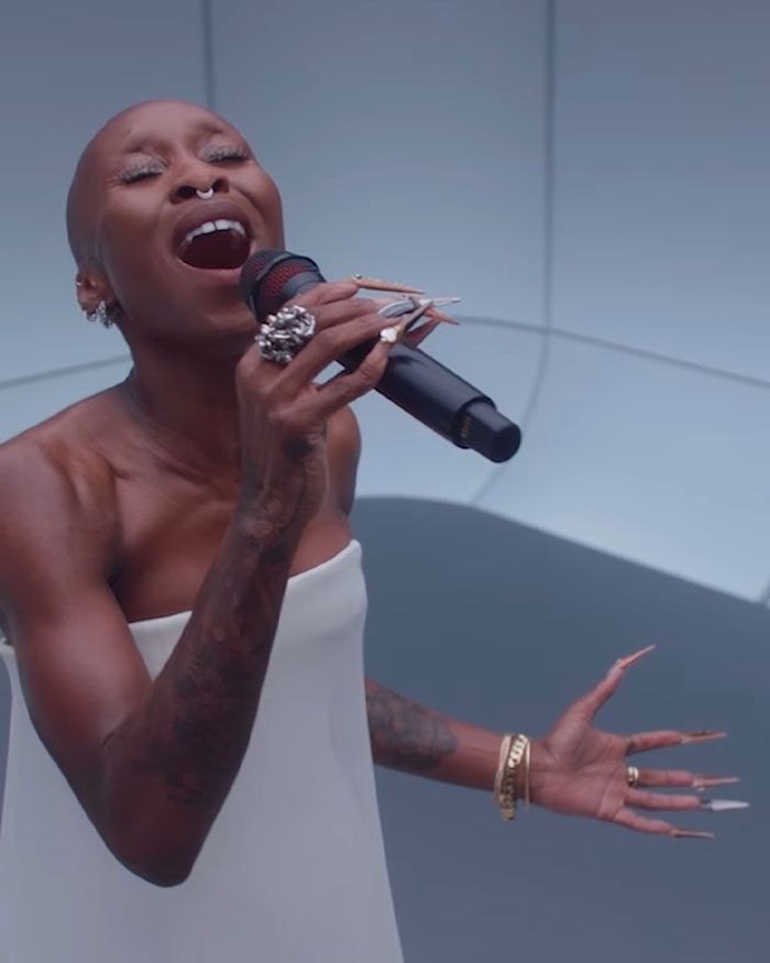 Cynthia Erivo singing passionately wearing a white dress, highlighting her oddly large hands and long nails in a performance.