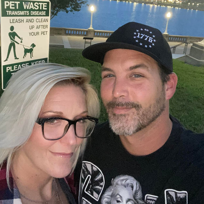 Man and woman posing outdoors near waterfront at dusk with pet waste sign in background during family gathering.