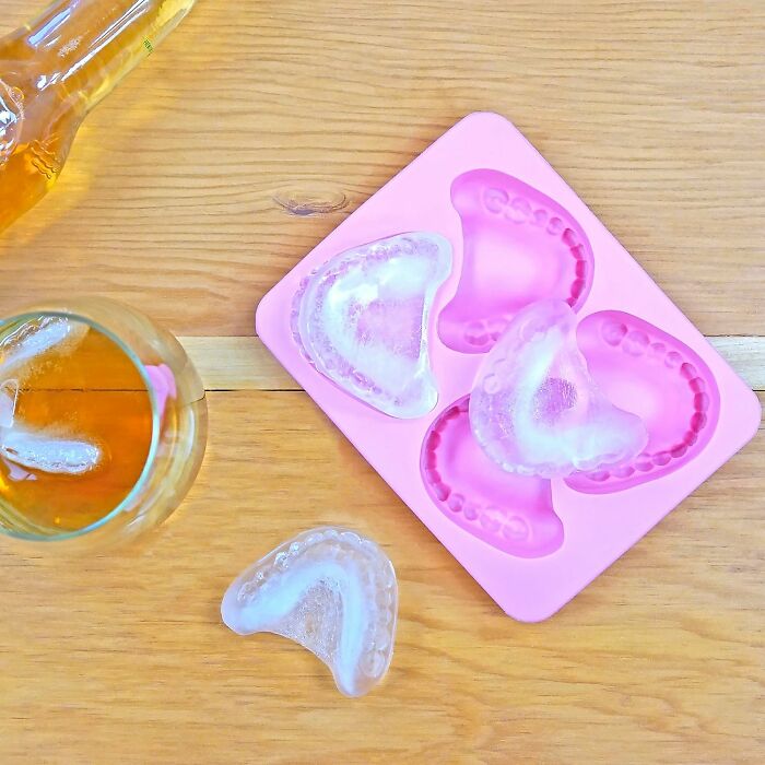 Ice cubes shaped like teeth made with a pink silicone mold, next to a glass of amber liquid on a wooden surface.