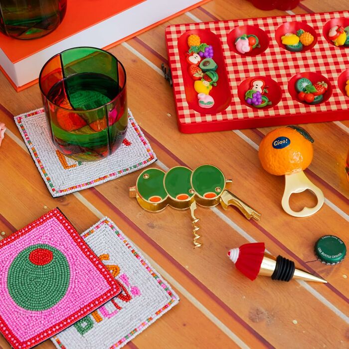 Colorful quirky gifts including a green glass, fruit-themed bottle opener, and beaded coasters on a wooden table.
