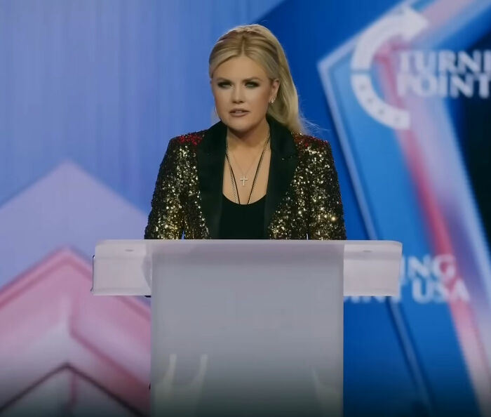 Erika Kirk speaking at TPUSA event, wearing a black and gold jacket during the greatest Freudian slip moment.