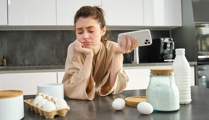 Woman at kitchen counter looking frustrated holding phone, dealing with picky eater and allergy concerns.