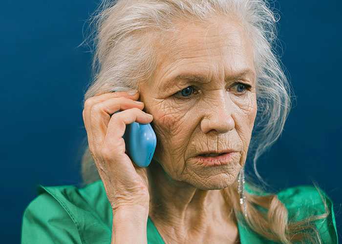 Elderly woman in green shirt holding a blue phone to her ear, looking shocked by what her MIL just said.