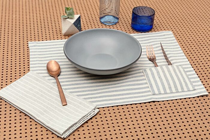 Minimalist table setting with striped placemat, gray bowl, copper utensils, and blue drinking glasses as unique gift ideas.