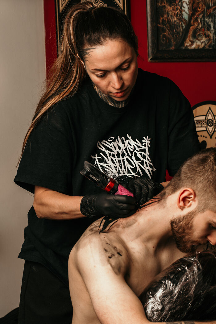 Female tattoo artist from Sacred Gold creating detailed neck tattoo on male client in studio setting.