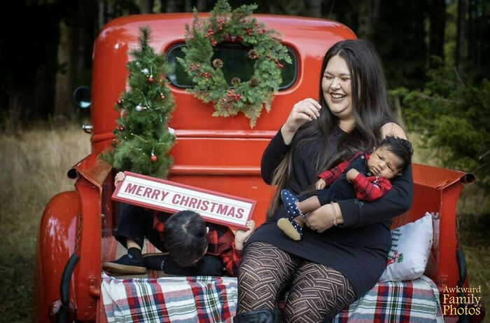 A woman holding a baby laughs while a child hides behind a Merry Christmas sign in an awkward family Christmas photo.