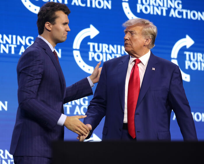Charlie Kirk and Donald Trump shaking hands onstage at a Turning Point Action event with blue branded background.
