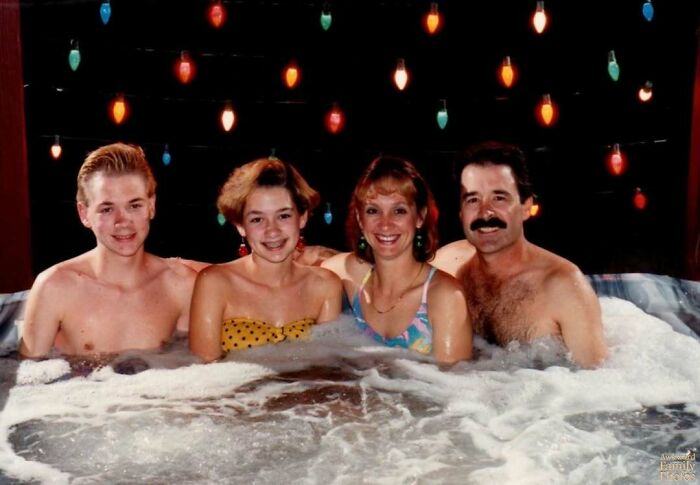 Awkward family Christmas photo of four people in a hot tub with festive colorful lights in the background.