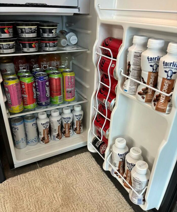 Mini dude fridge filled with assorted drinks including soda cans, sparkling water, and protein shakes on carpeted floor.