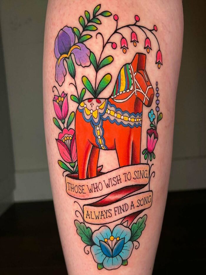 Colorful wholesome tattoo of a traditional horse surrounded by flowers and a banner with a meaningful quote on the leg.