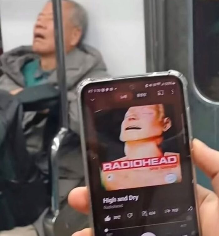 Person holding phone showing Radiohead album cover with a sleeping man in background, related to Know Your Meme.