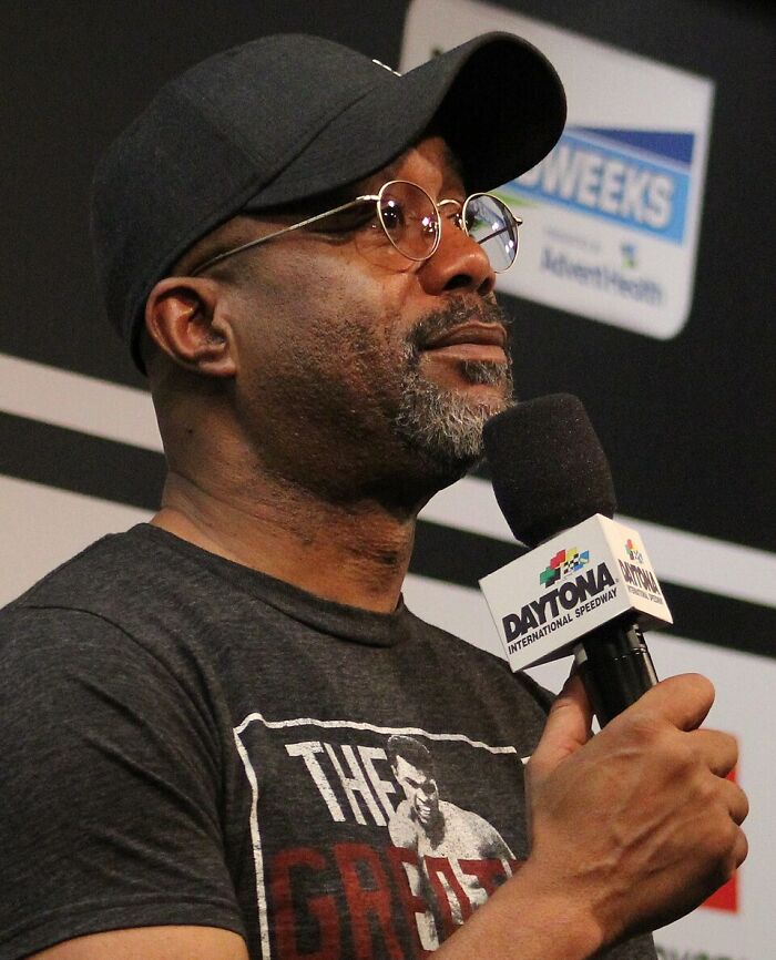 Man wearing glasses and cap speaking into a microphone at Daytona International Speedway event about once famous regular job.