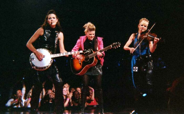 Three female musicians performing on stage with banjo, guitar, and violin, spotlighted in a cancel culture victims concert.