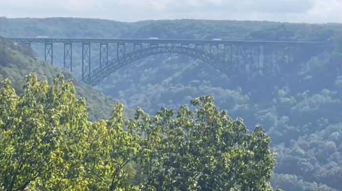 Steel arched bridge spanning a forested valley with hills in the background under a cloudy sky, urban legends theme.