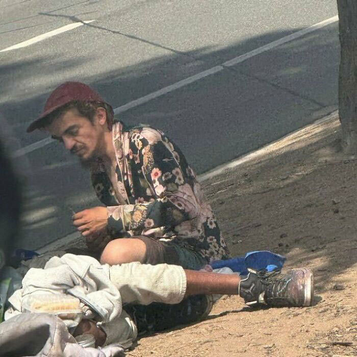 Man wearing a floral shirt and red cap sitting on sidewalk near homeless person, highlighting Tylor Chase homelessness concern.