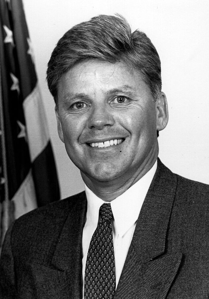 Smiling man in a suit and tie with an American flag in the background, representing cancel culture victims celebrities.