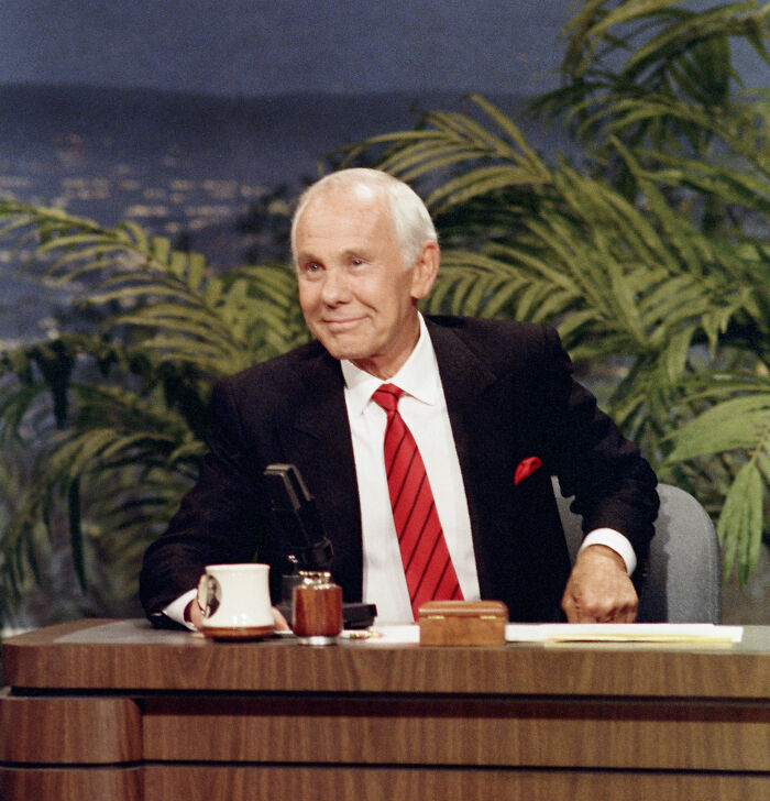 Johnny Carson hosting the Tonight Show, seated at the desk with a microphone and plants in the background.
